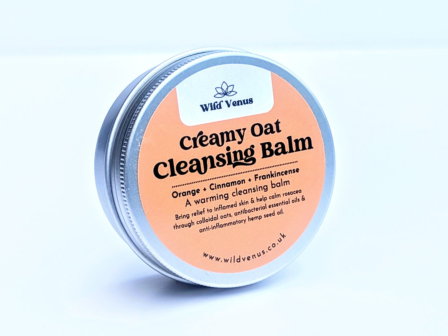 A tin of Creamy Oat Cleansing Balm on its side and slightly tilted away from the camera, showing the top of the tin with the label showing. The product is against a plain white background.