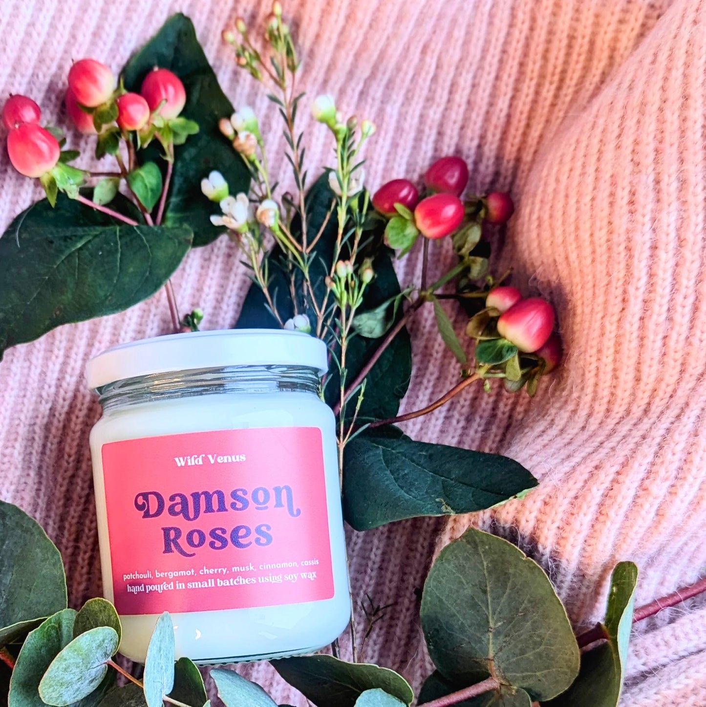 A damson roses soy candle is laying down on a pink wollen surface. The rose scented candle is surrounded by foliage and berries. The soy candle is off center to the left of the square frame.