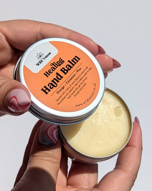 A pair of hands are shown holding the Healing hand Balm. The left hand holds the lid and shows the label, the right hand is holding the bottom half od the product which contains the hand balm.