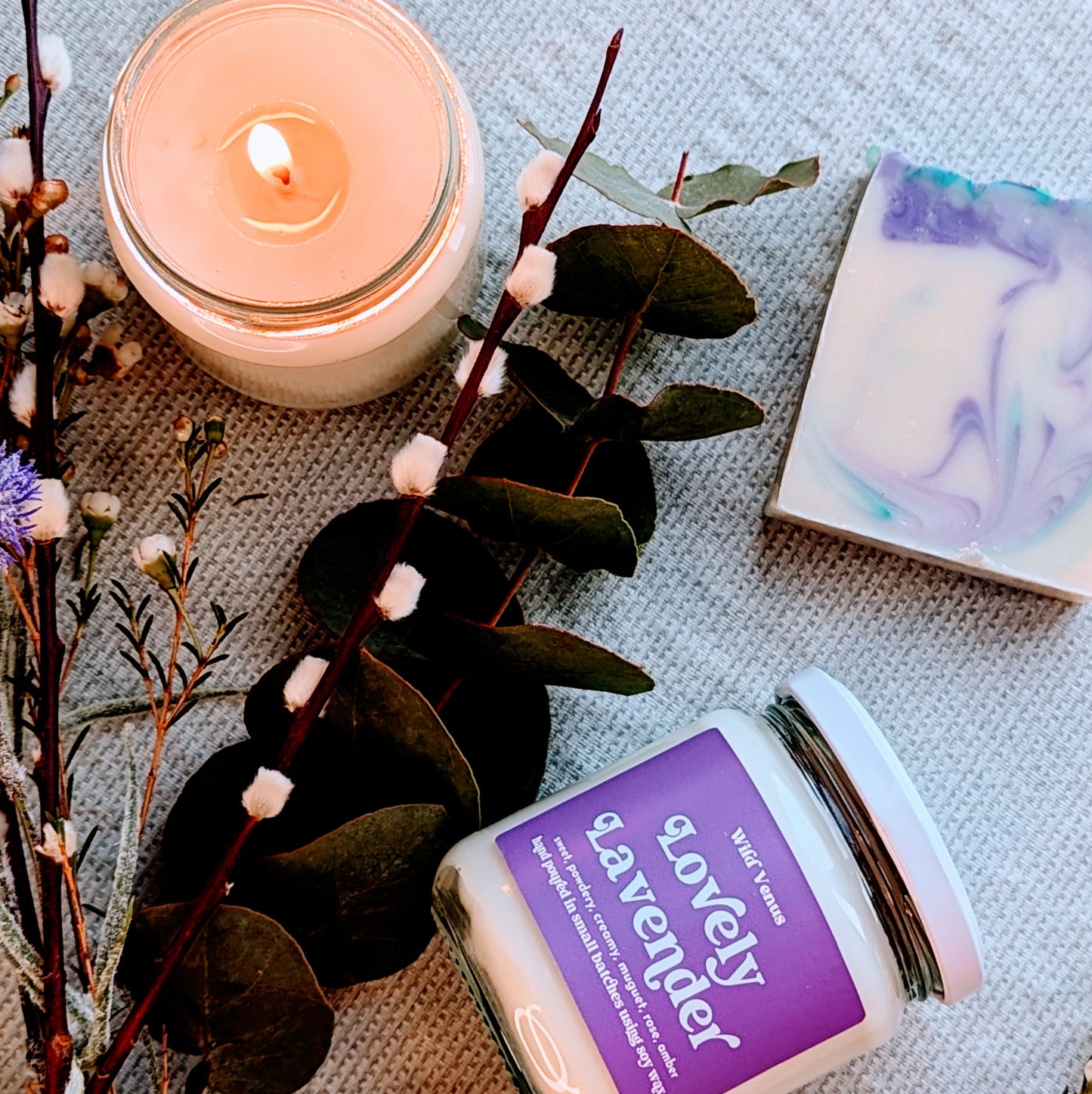 A flay lay photo view from above of arranged foliage including lavender and eucalyptus. Nestled in the greenery is a lit soy candle and a lovely lavender candle laying on its side showing its label. There is also a lavender soap with green and purple swirls.