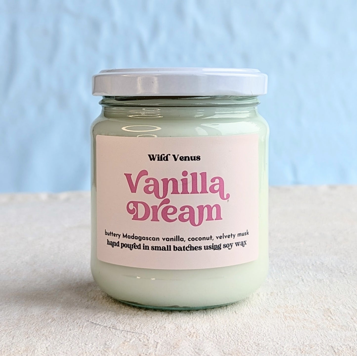A Vanilla dream soy candle is on a white surface and against a plain light blue background. The vanilla candle has its lid on.
