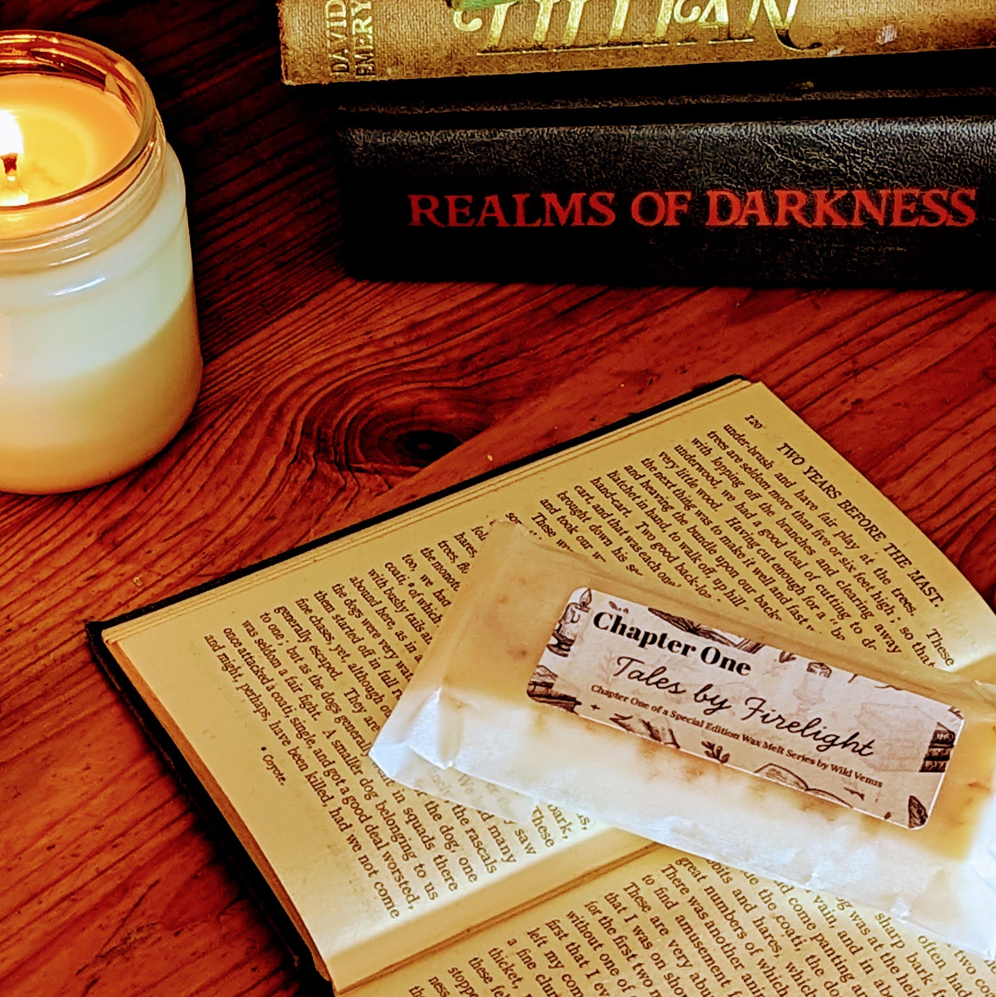 A soy wax melt is on top of an open book, candles flicker beside it and there is a stack of books, ivy peeks I the background. 