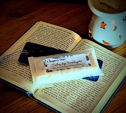 There is an open book with a bookmark and a wax melt on top. A wax warmer is beside the book and the photo has a rich sepia tone. 
