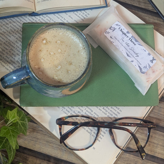 There is a blue mug in the centre of the picture, you can see the mug from above, it's almost full with white frothy liquid. Beside the mug is a wax melt, there is also a pair of glasses. These items are on top of books. Some books are open and some are closed. 