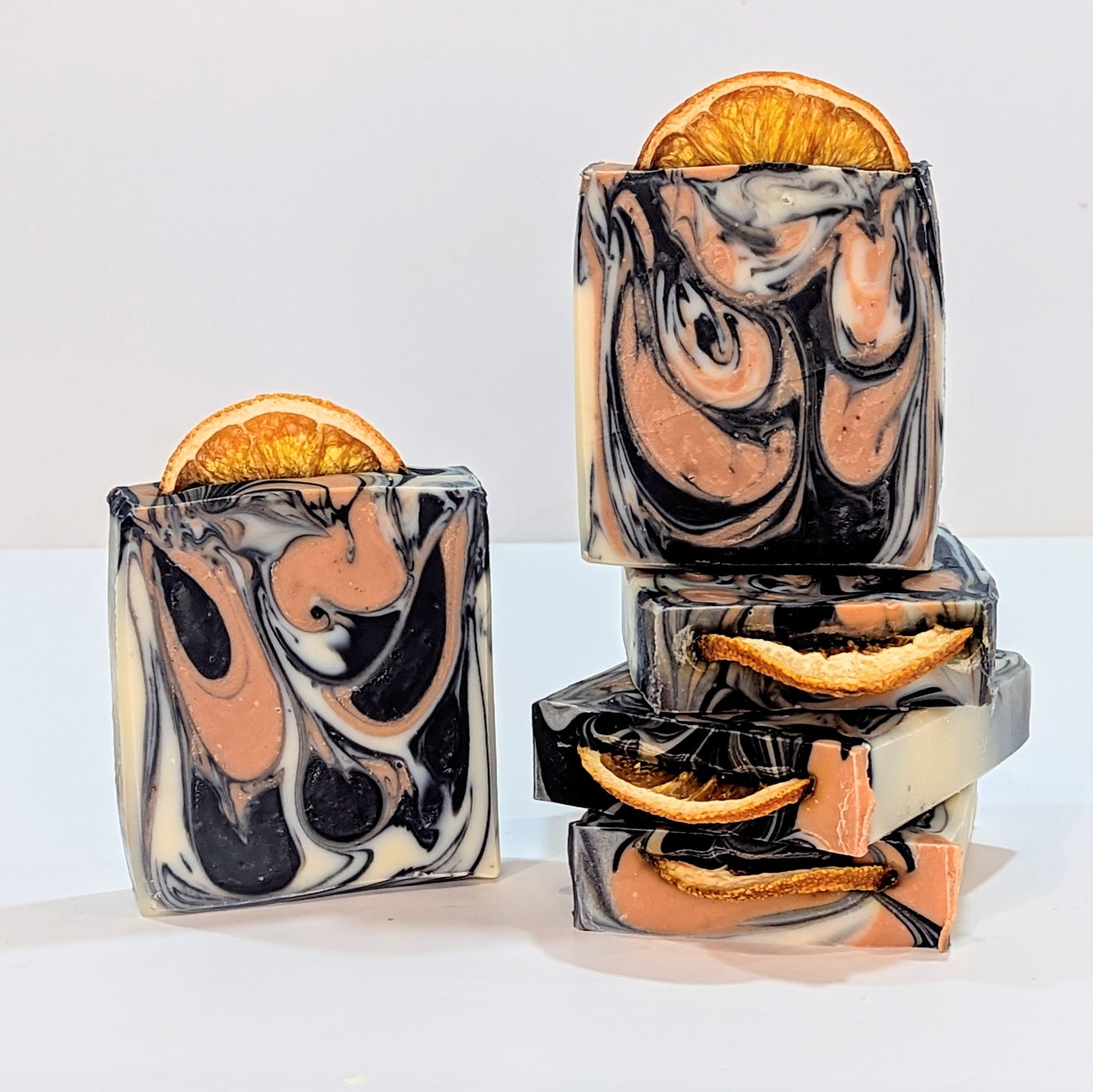 There are three bars of soap laying flat. Standing upright a soap is beside them and another soap is on top of them. The soaps are swirly with beetroot powder and charcoal. A slice of orange is on the top.  The soaps are called Clear Complexion. 