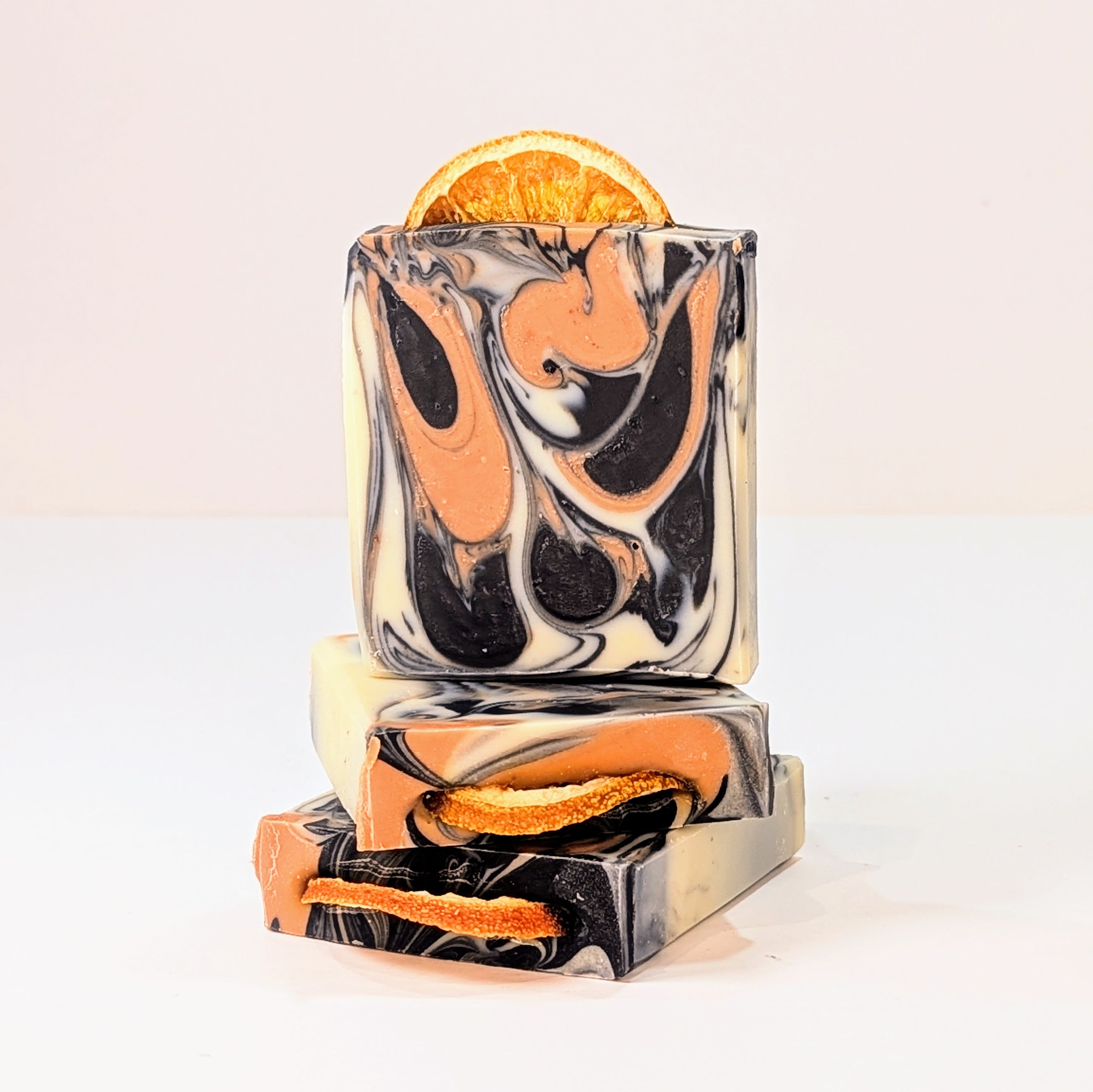 In front of a white background, there are two bars of soap are on the side. Another soap is on top of them. The soaps are swirly with beetroot powder and charcoal. A slice of orange is on the top. 