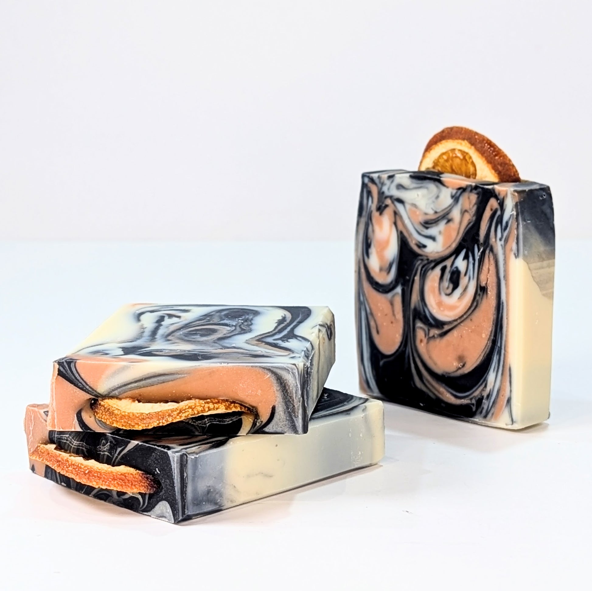 Two bars of soap are on the side and at an angle. Another soap is next to them. The soaps are swirly with beetroot powder and charcoal. A slice of orange is on the top of each soap bar.