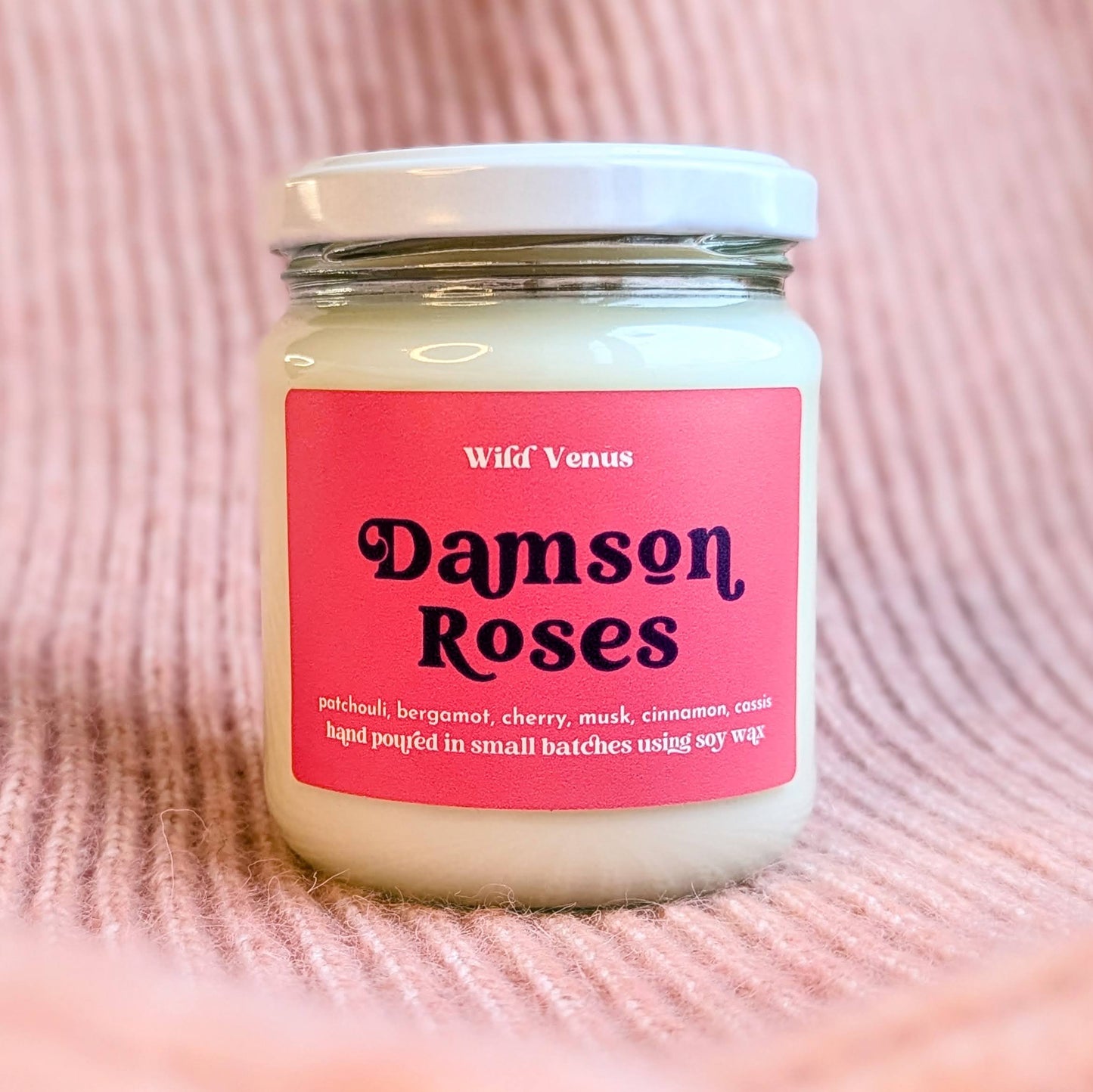 A soy candle is shown against a pink woollen and ribbed effect surface. The container candle has its lid on and is called Damson Roses. The candle has a red label with descriptive retro font. 