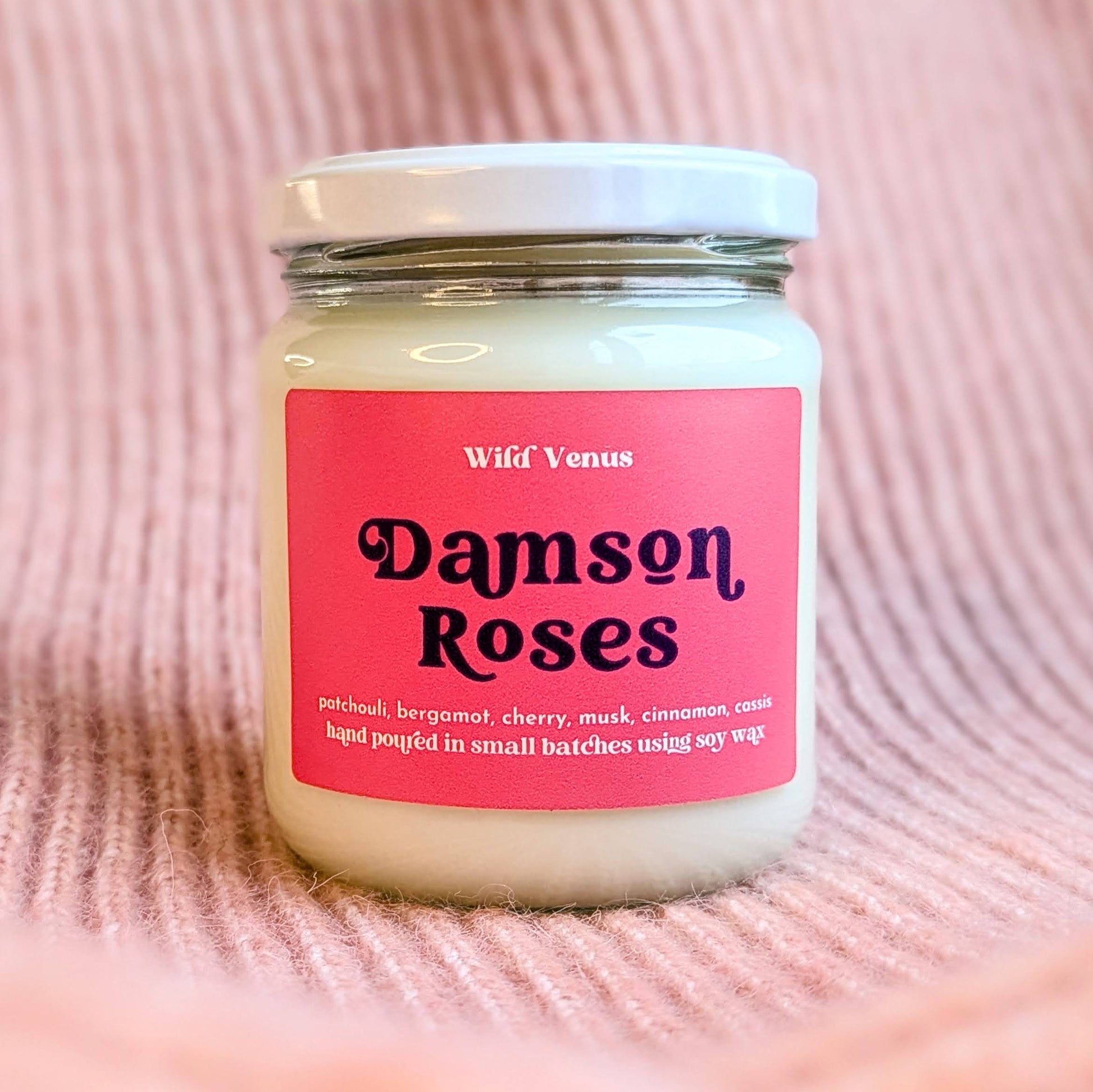 A soy candle is shown against a pink woollen and ribbed effect surface. The container candle has its lid on and is called Damson Roses. The candle has a red label with descriptive retro font. 