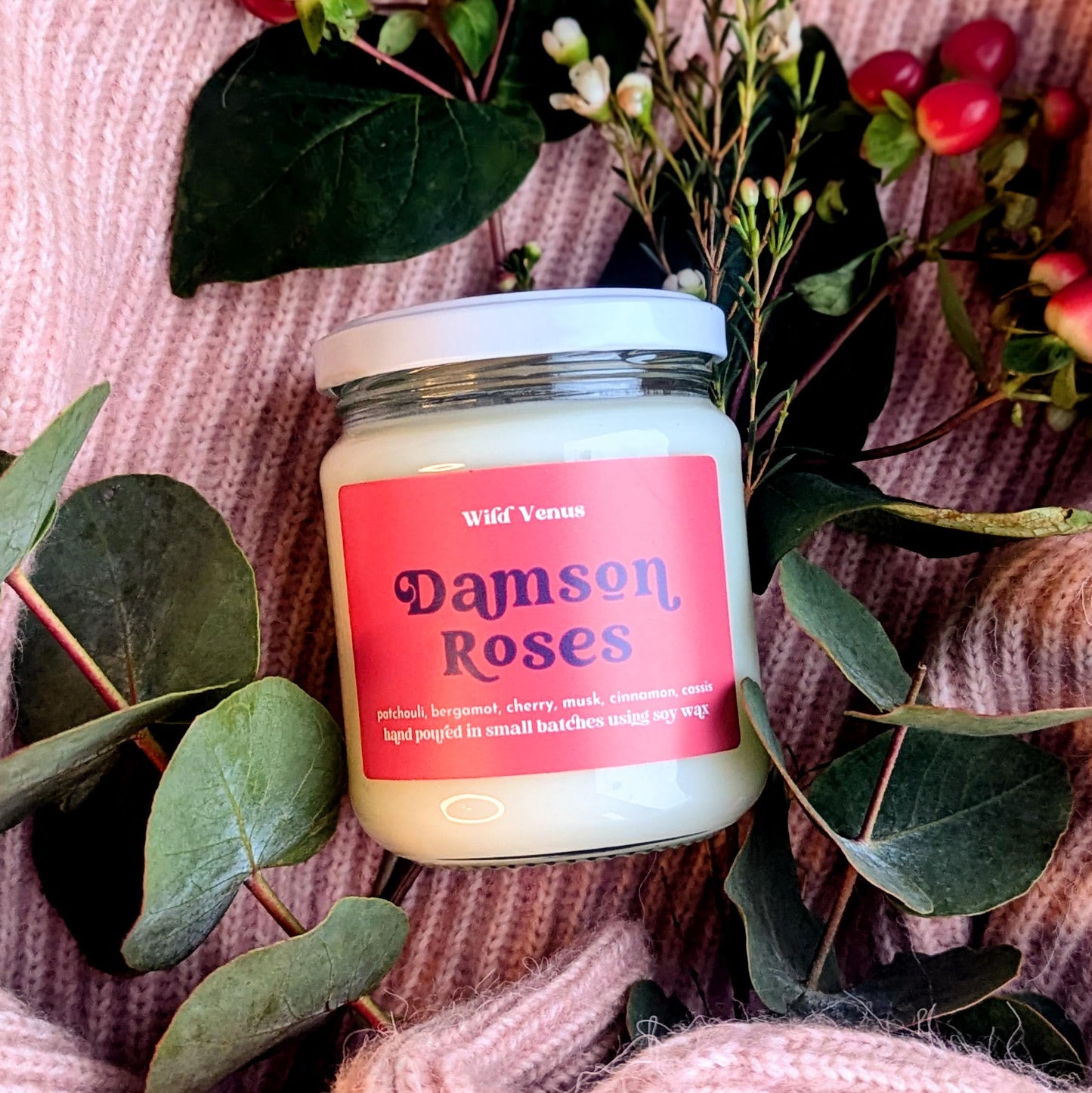 The Damson Roses handmade soy candle is shown against a pink background. The candle is in a flat lay position and central in the square photo. There are eucalyptus leaves around the candle and a few berries. 