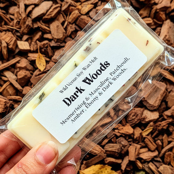 Hand holding a wax melt package labeled 'Dark Woods' against a wood chip background
