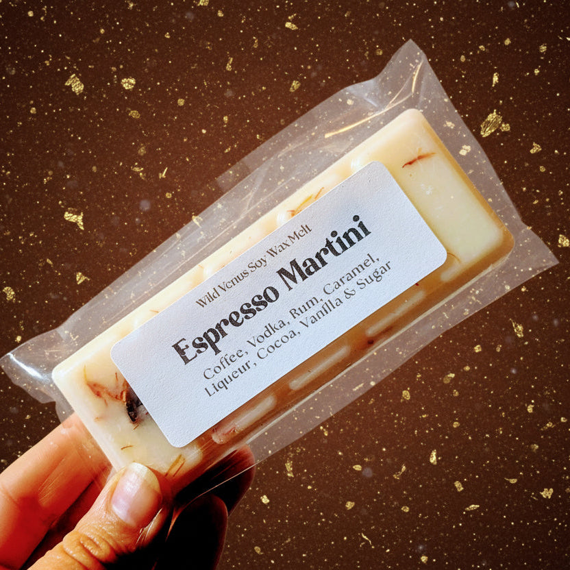 Hand holds an espresso martini scented wax melt against a brown and gold background. 