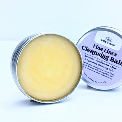 A tin of Fine Lines Cleansing Balm. The tin is open and lid placed slightly behind the open tin. This tin is on its side and front on so you can see the whole label on the tin lid. The product is shot against a plain white background. 