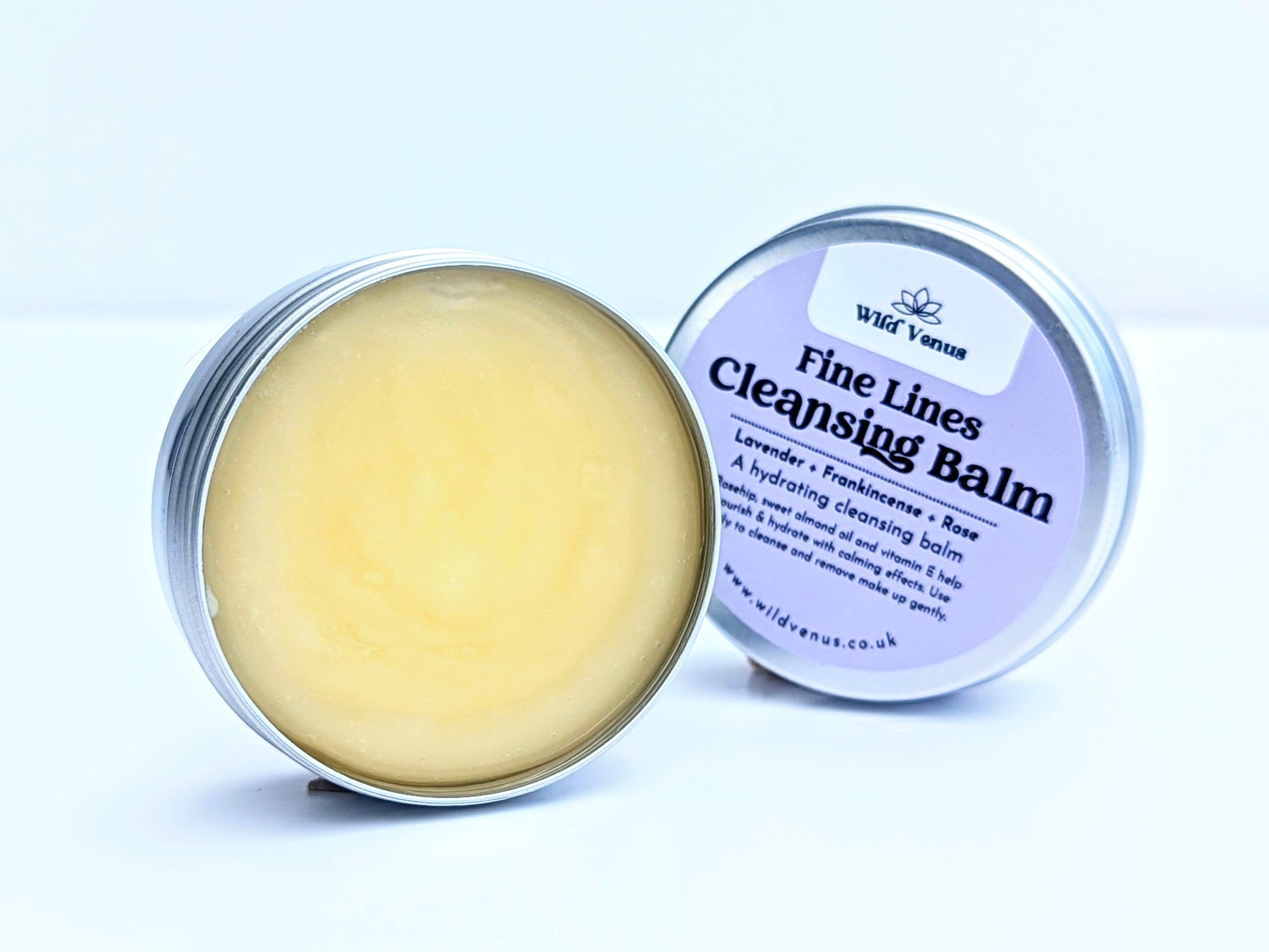 A tin of Fine Lines Cleansing Balm. The tin is open and lid placed slightly behind the open tin. This tin is on its side and front on so you can see the whole label on the tin lid. The product is shot against a plain white background. 