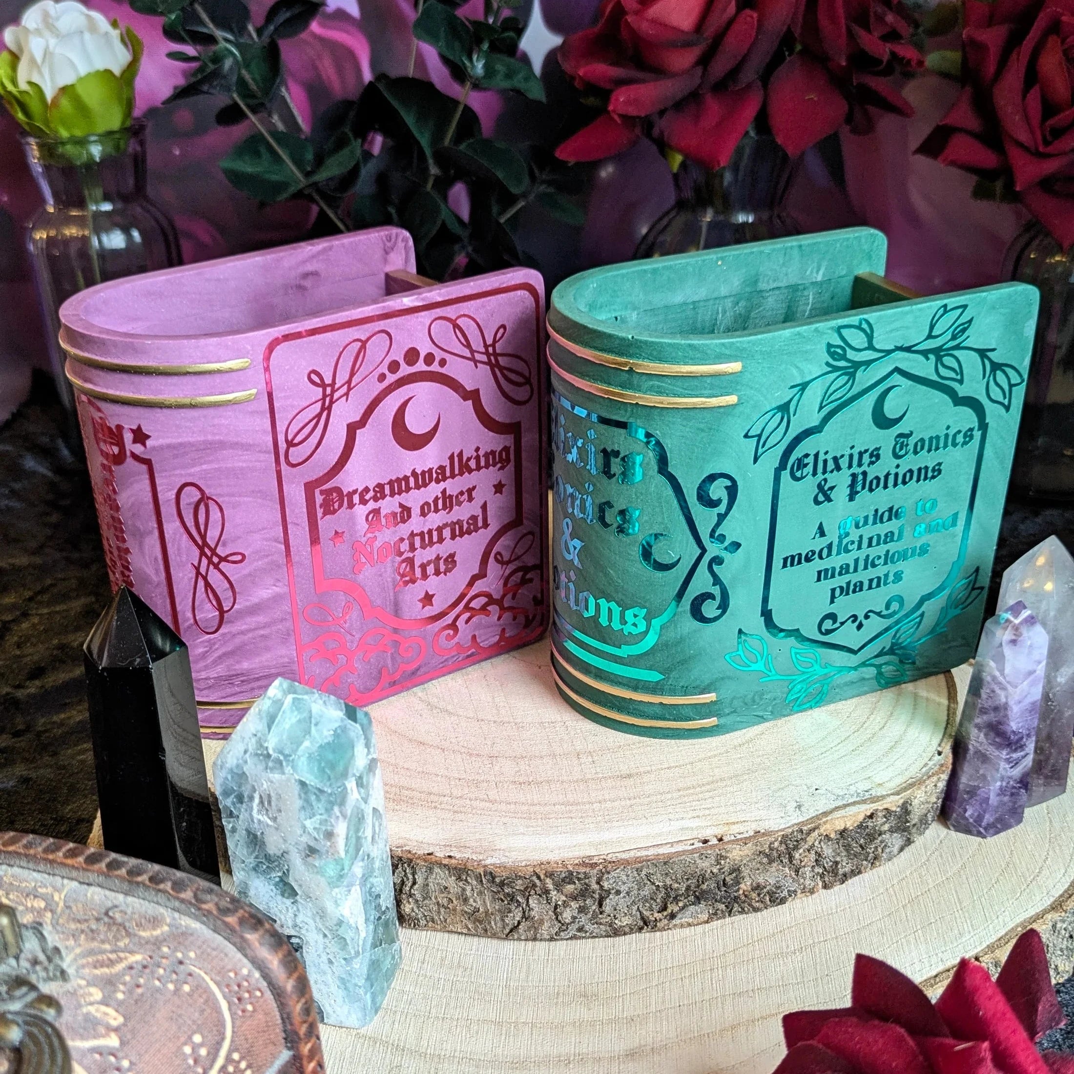 Two decorative jesmonite bookmark containers with fairy-themed designs on a wooden surface with flowers and crystals.