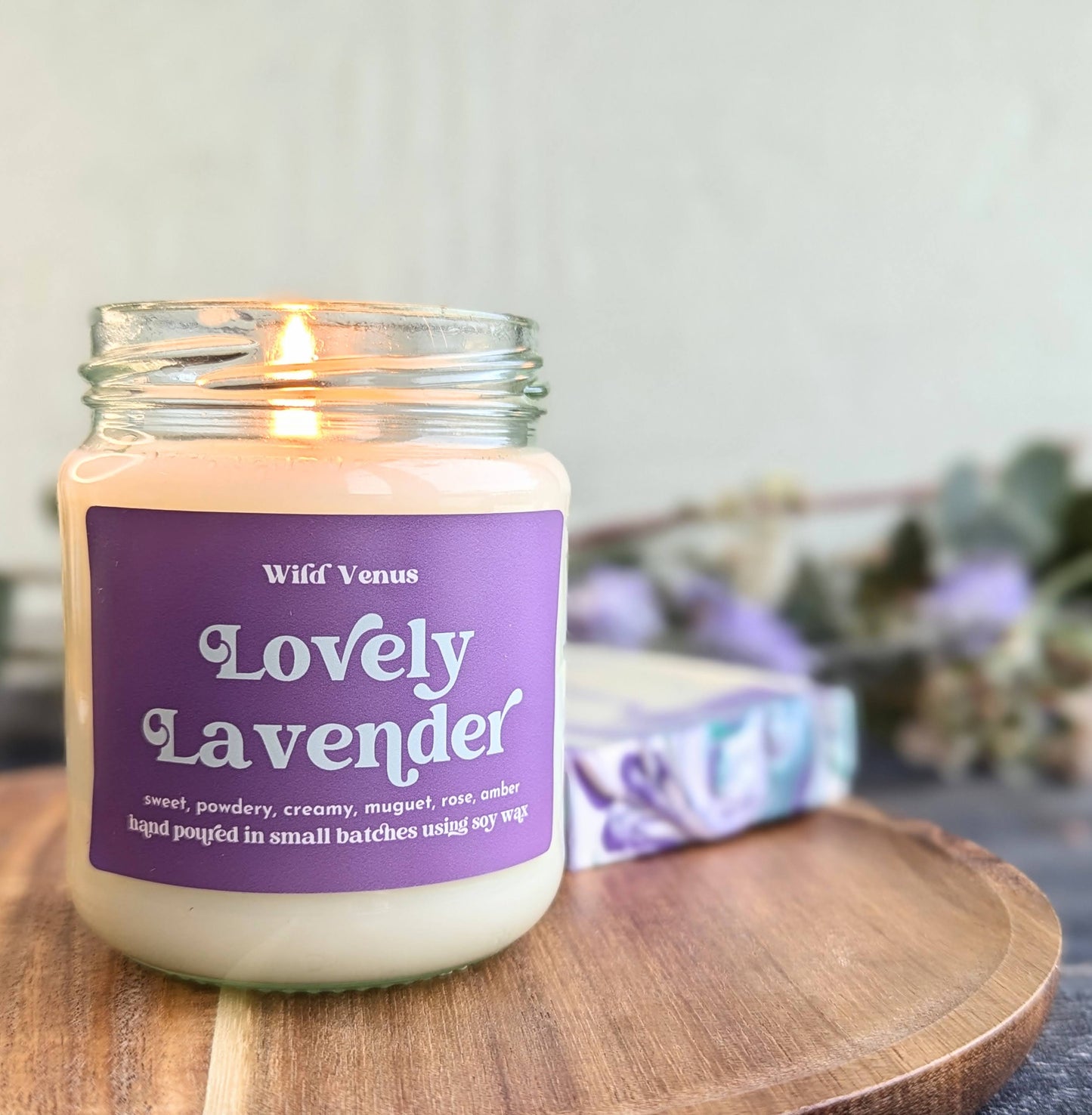A Lovely Lavender scented soy candle is lit and placed on a small wooden board in in the left of the frame. Behind the soy candle is a lavender soap and some foliage. 