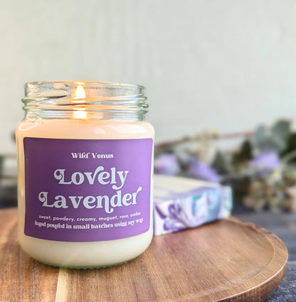 A Lovely Lavender scented soy candle is lit and placed on a small wooden board in in the left of the frame. Behind the soy candle is a lavender soap and some foliage. 
