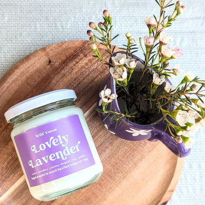 The lovely lavender scented candle is on a small wooden circular chopping board, laying on its side with its label visible. The view is from above and beside the soy candle is a small decorative purple jug filled with daisys. 