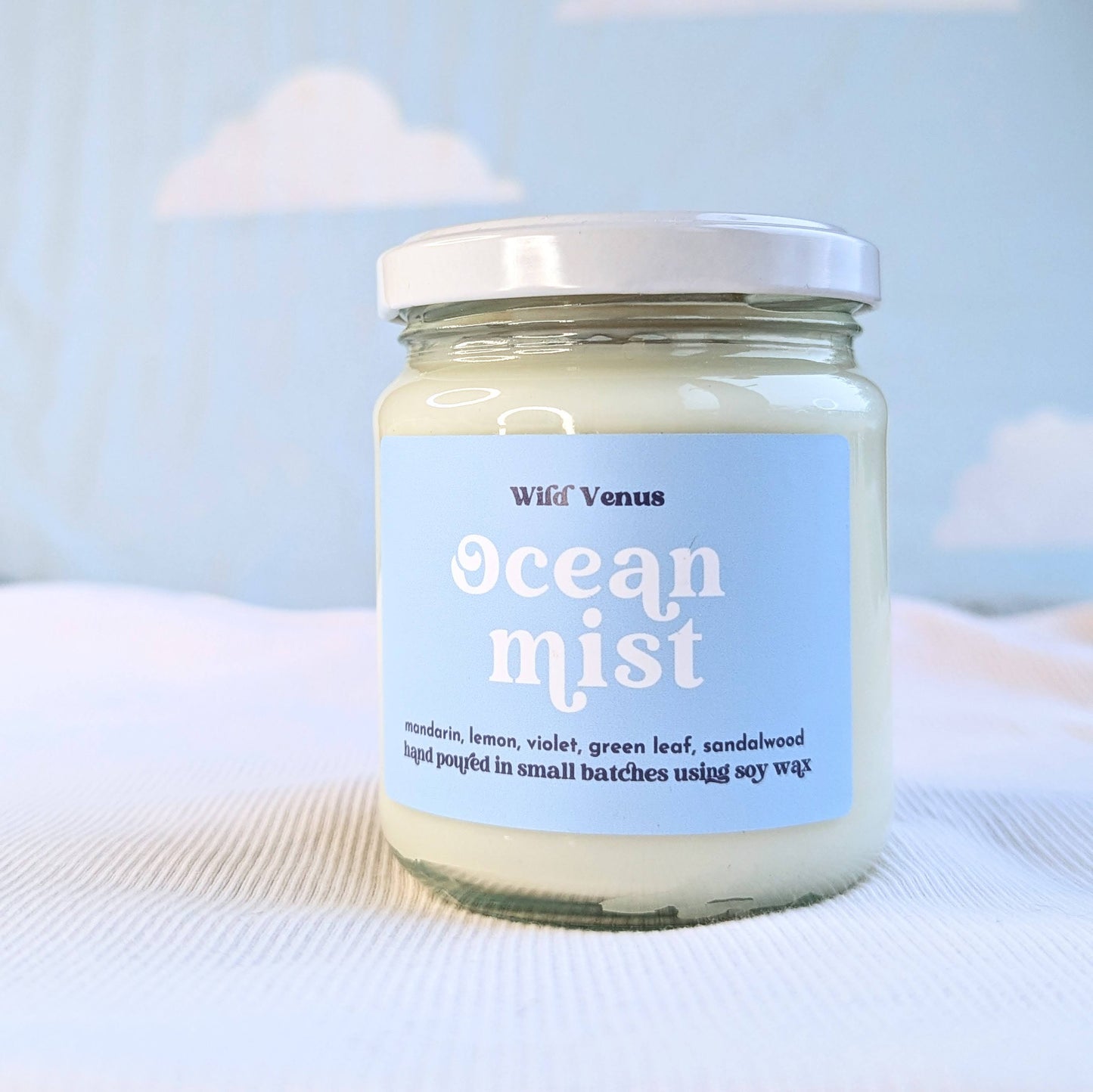 A soy container candle is on a soft white surface against a blue background. The candle is called Ocean Mist and it has a lid.