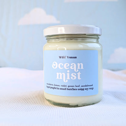 A soy container candle is on a soft white surface against a blue background. The candle is called Ocean Mist and it has a lid.