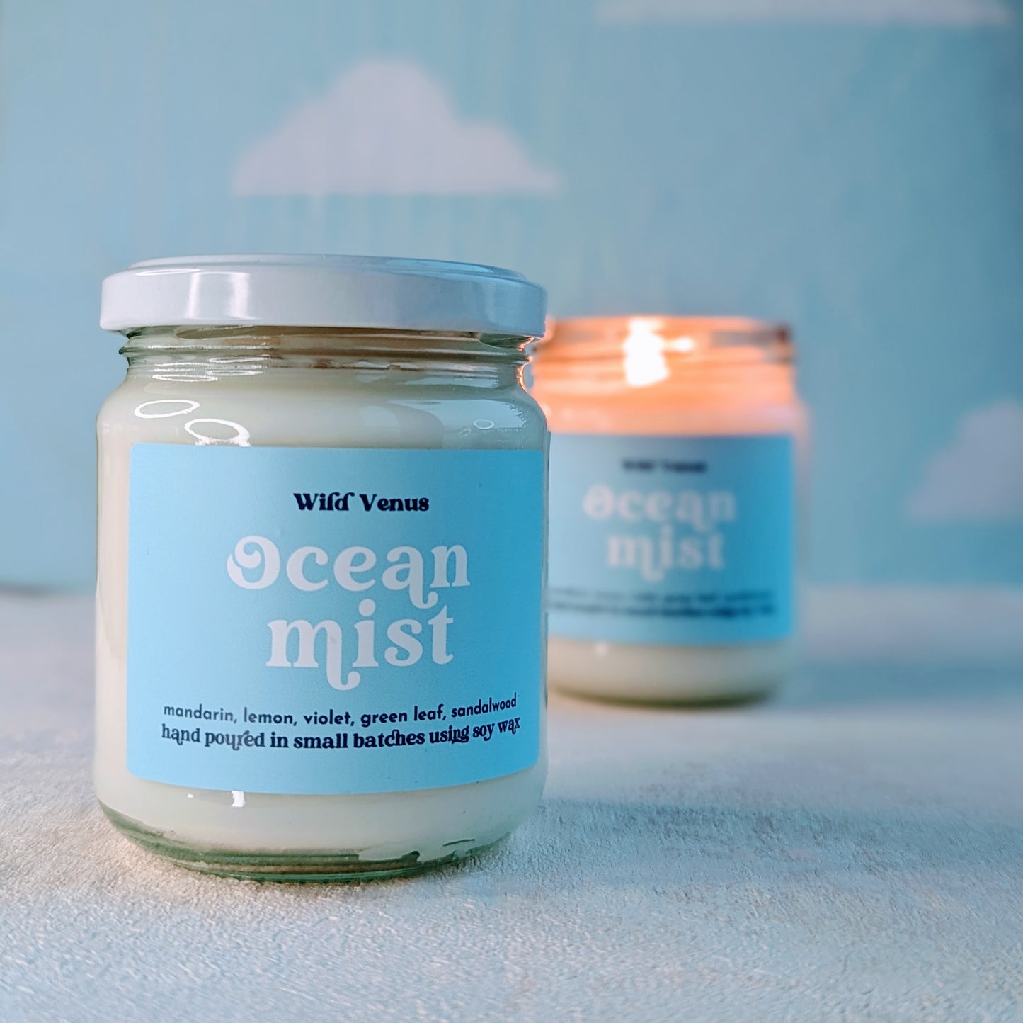 An Ocean Mist scented candle is in front of a lit Ocean Mist soy candle. They are both on a soft white surface and against a blue background with clouds. 