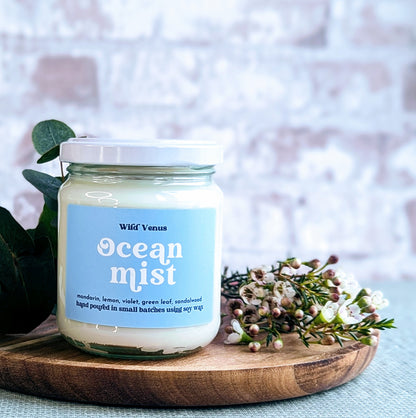 A soy container candle is on a wooden chopping board and against a light bright background. The candle is called Ocean Mist and beside it are some eucalyptus leaves and some small flowers. 
