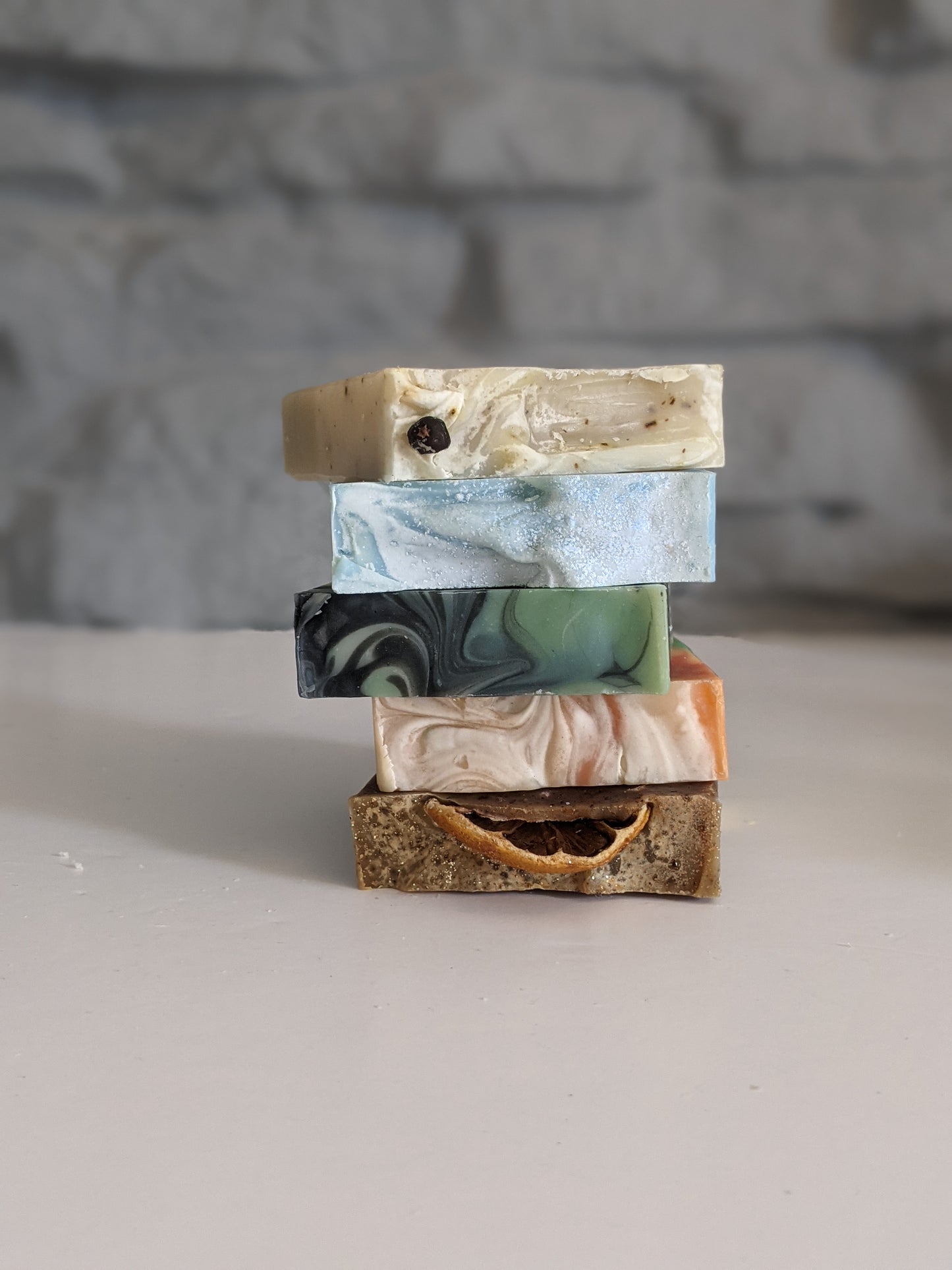 A stack of 5 different soaps on a white surface, against a grey brick background.