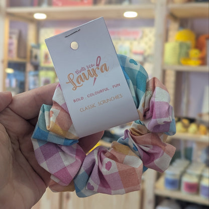 A hand holds a pastel plaid fabric scrunchie with a white paper hangtag that reads 'Sandy's' and 'Bold Coloured Fun Classic Scrunches' in a shop with shelves of colorful items in the background.