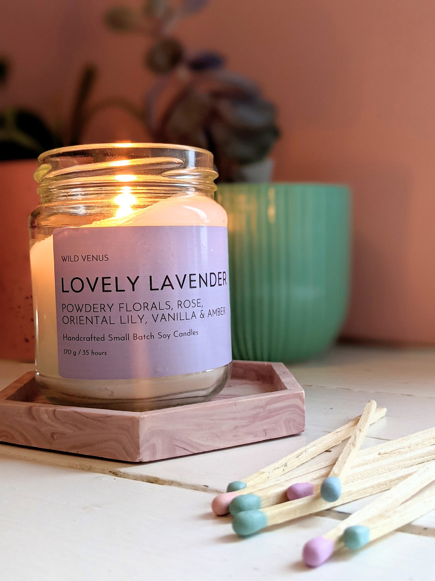 A lit Lovely Lavender candle on a pink coaster and some pastel matches in the foreground.