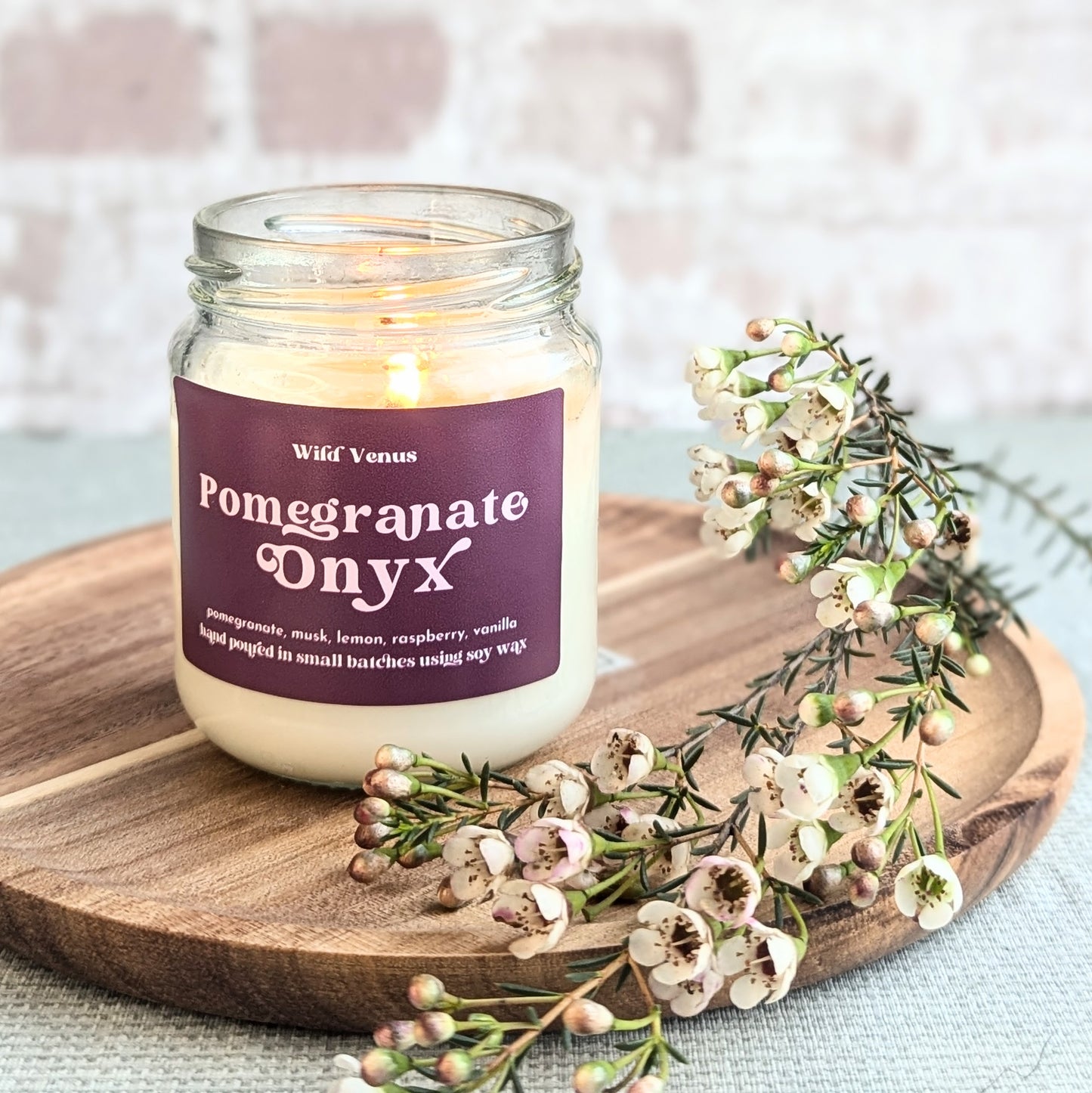 A lit soy candle is on a wooden chopping board with a sprig of flowers beside it. The candle is pomegranate scented and called pomegranate onyx.