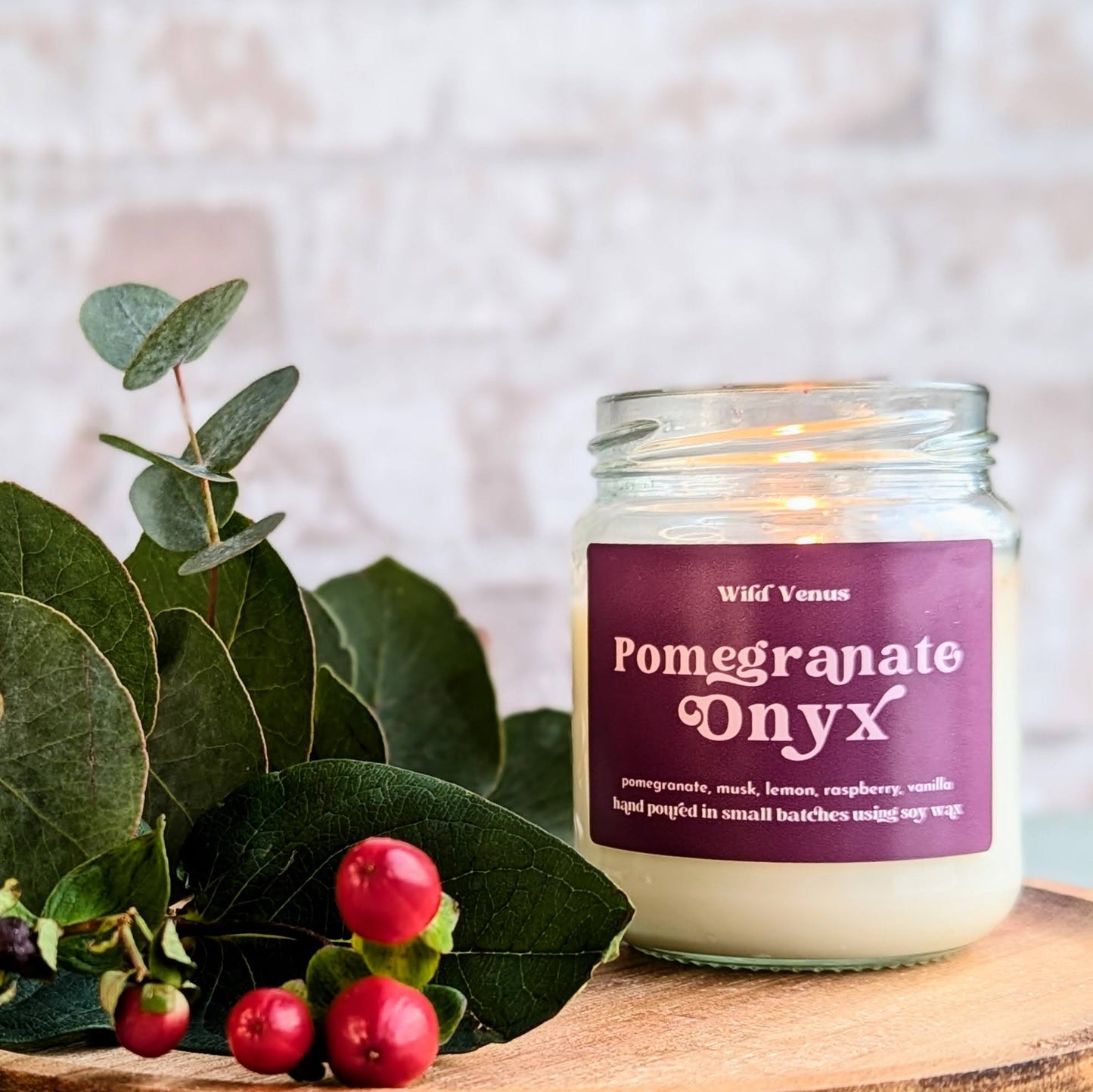 A lit pomegranate soy candle is on a wooden board beside some berries and foliage. The scene is against a stone background.
