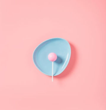 A pink lolly is on a blue dish, against a pale pink background. 