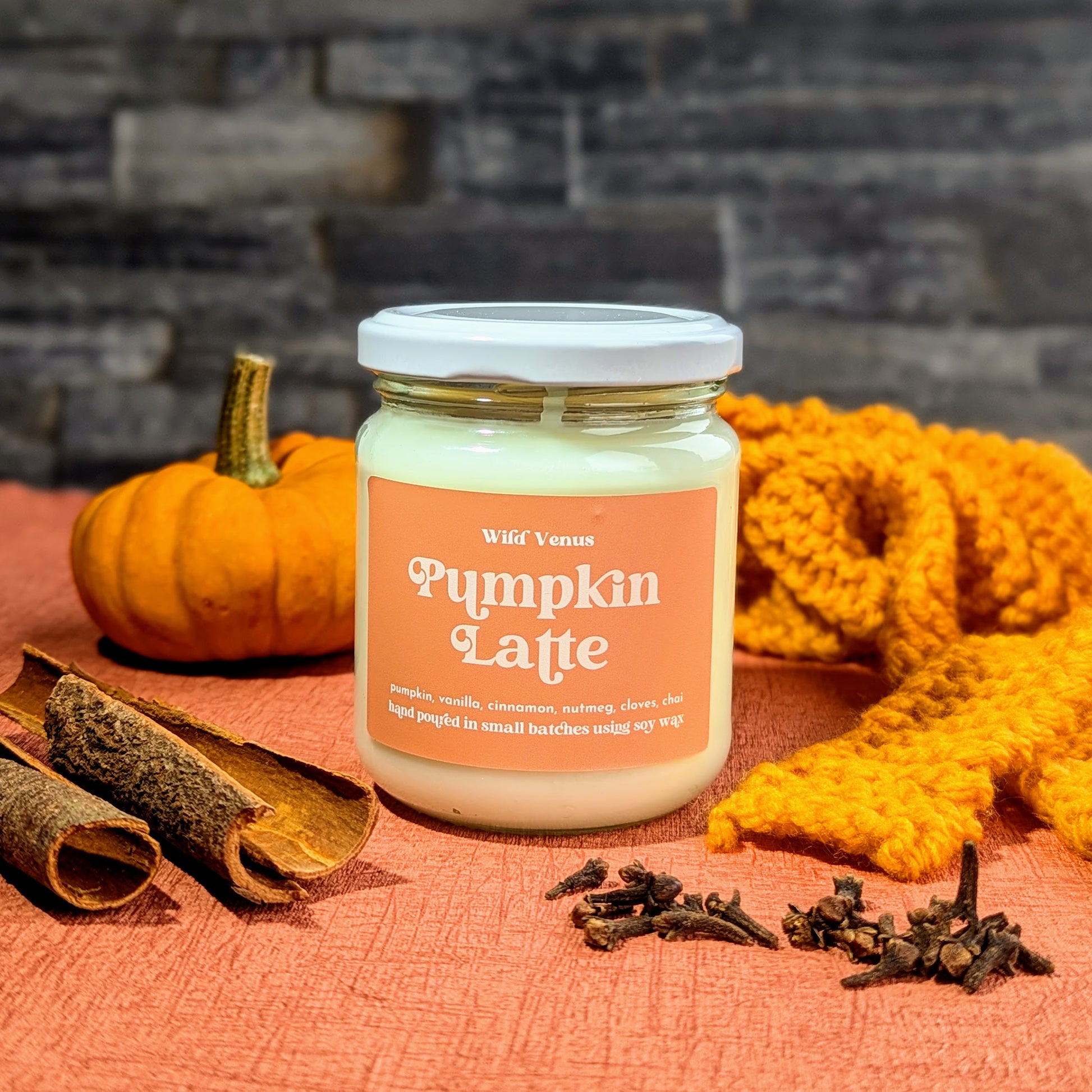 A candle with the Pumpkin candle label on, it is on an orange surface with a slate brick wall background. In the foreground is cinnamon sticks, cloves, a mini pumpkin and an orange scarf.