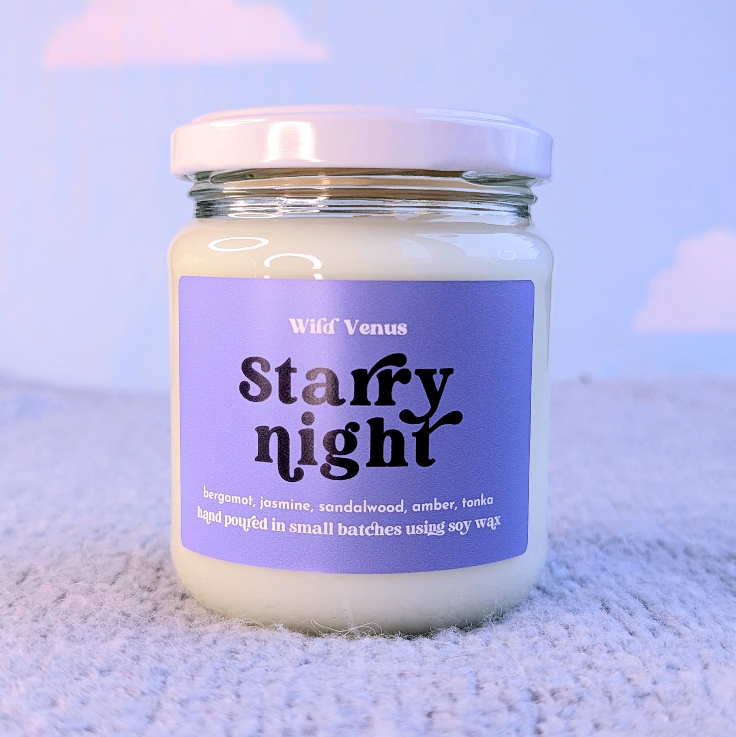 A starry night soy candle is on a light blue surface and against a light blue background with soft clouds. The soy container candle is shown with the lid on.