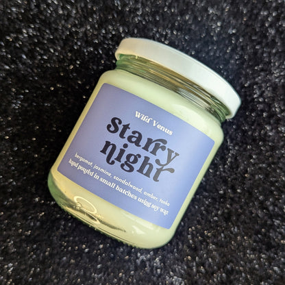The starry night soy candle is shown on its side with its label facing the viewer. The candle has been placed on a soft, black and sparkly surface. 