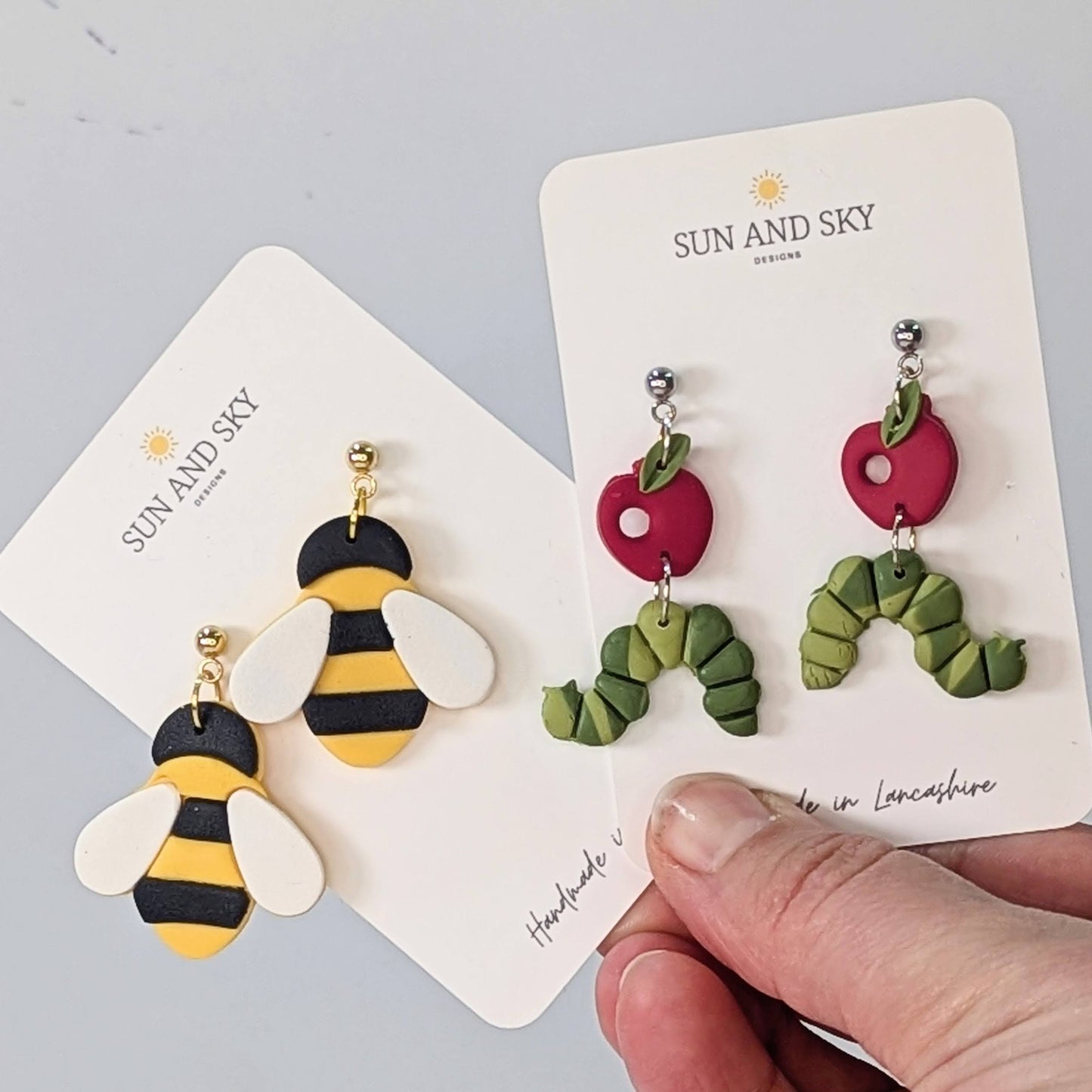 Holding two pairs of sun and sky earrings including bee earrings and caterpillar earrings. 