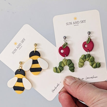 Holding two pairs of sun and sky earrings including bee earrings and caterpillar earrings. 