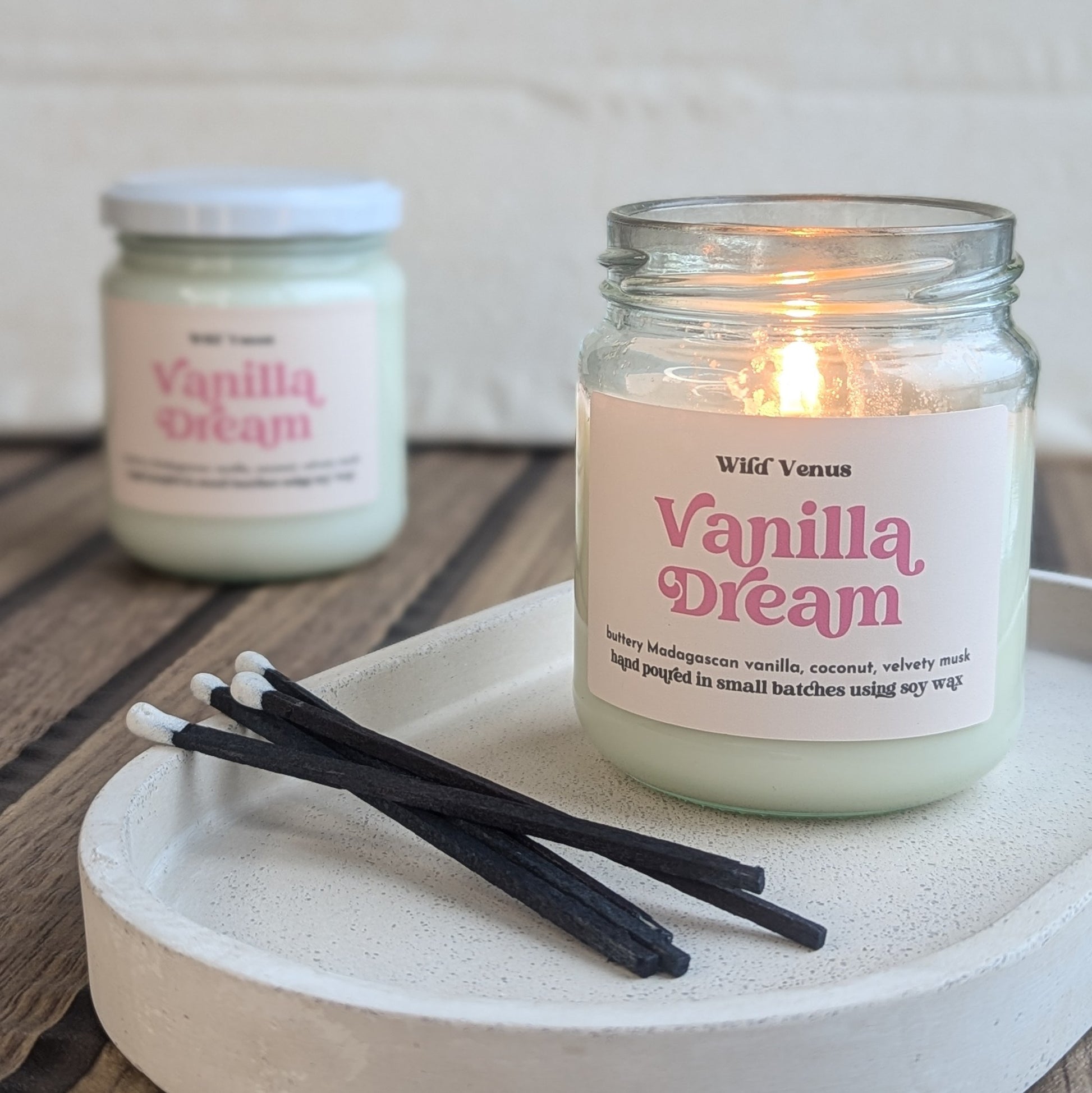 Two soy candles are shown in the picture. They are both scented in vanilla and are called 'Vanilla dream'. A candle which is closed is lit and the farthest one has its lid on. The lit soy candle is placed on a small stone trinket tray and there are some black matches beside it. 