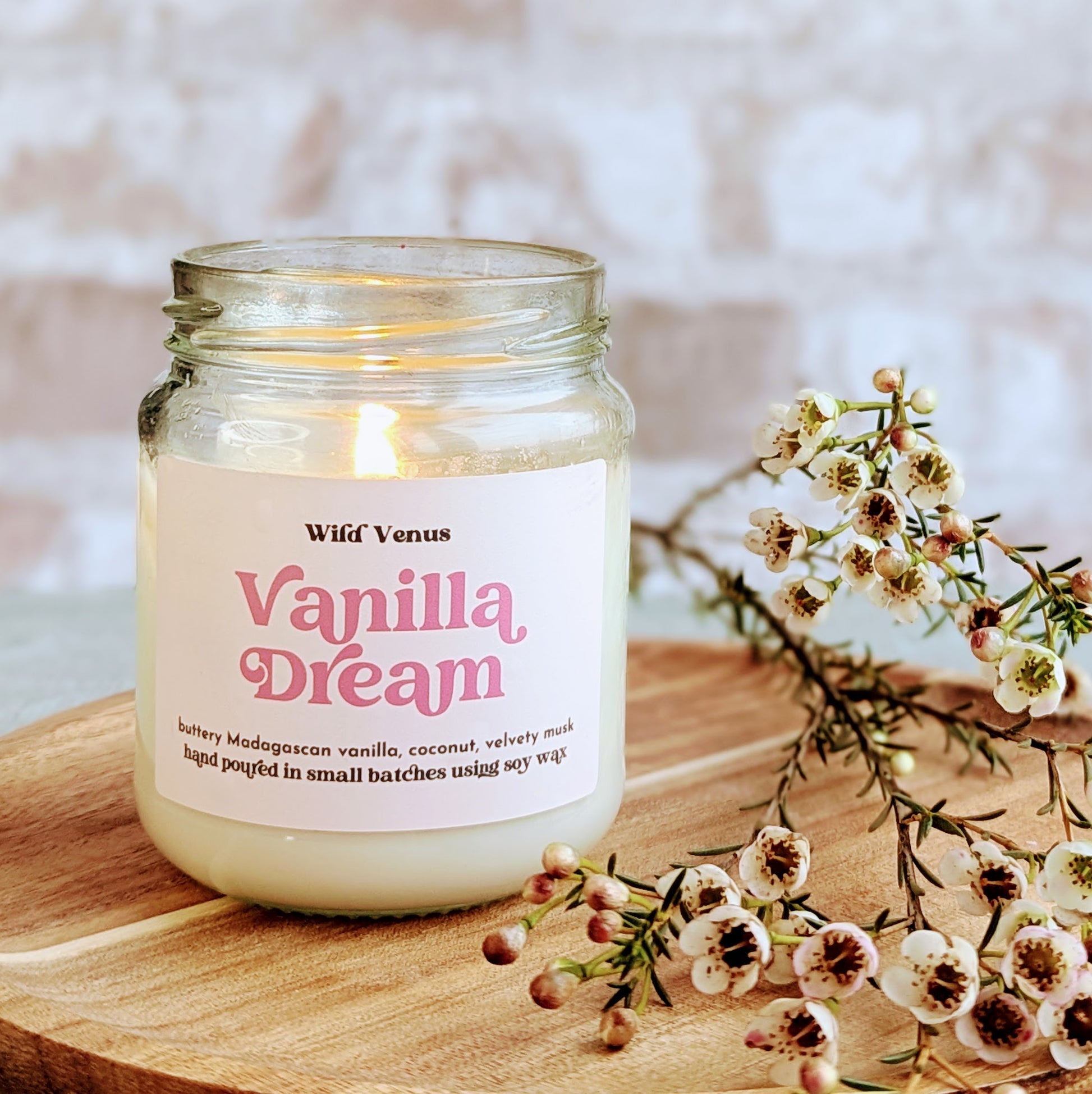 A lit soy candle is placed on a small wooden chopping board against a light brick background. The candle is called Vanilla Dream and there are small dainty flowers placed beside it on the chopping board. 
