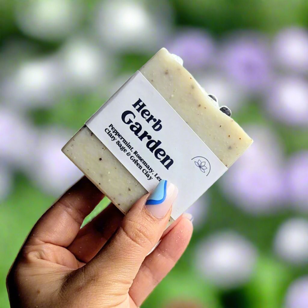 Holding a bar of herb garden soap up against a garden in the background. 