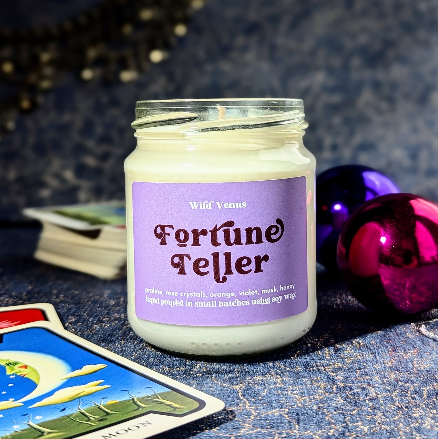 Fortune teller candle on a blue and gold table, there is a pack of tarot cards in the background and one card depicting the moon is visible in the foreground. There are some shiny baubles and some jewlery in the back.