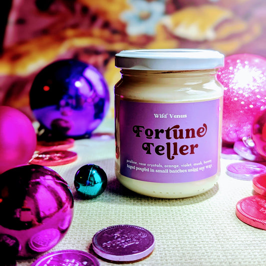 fortune teller soy wax candle is surrounded by shiny baubles, pink and purple coins. 