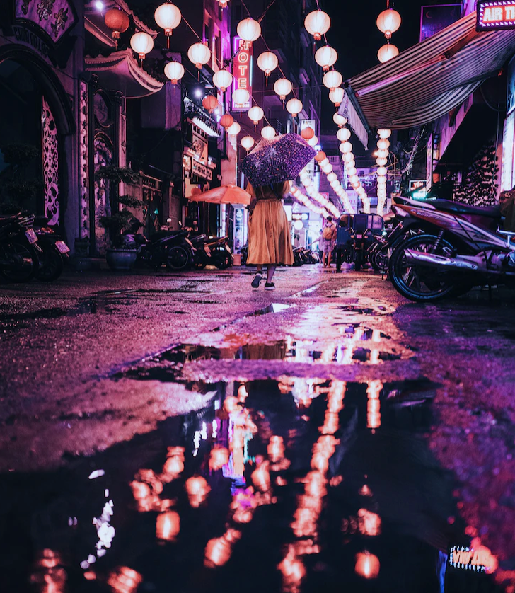 A lady walks away down a neon lit street. She's holding an umbrella and there are puddles in the ground. 